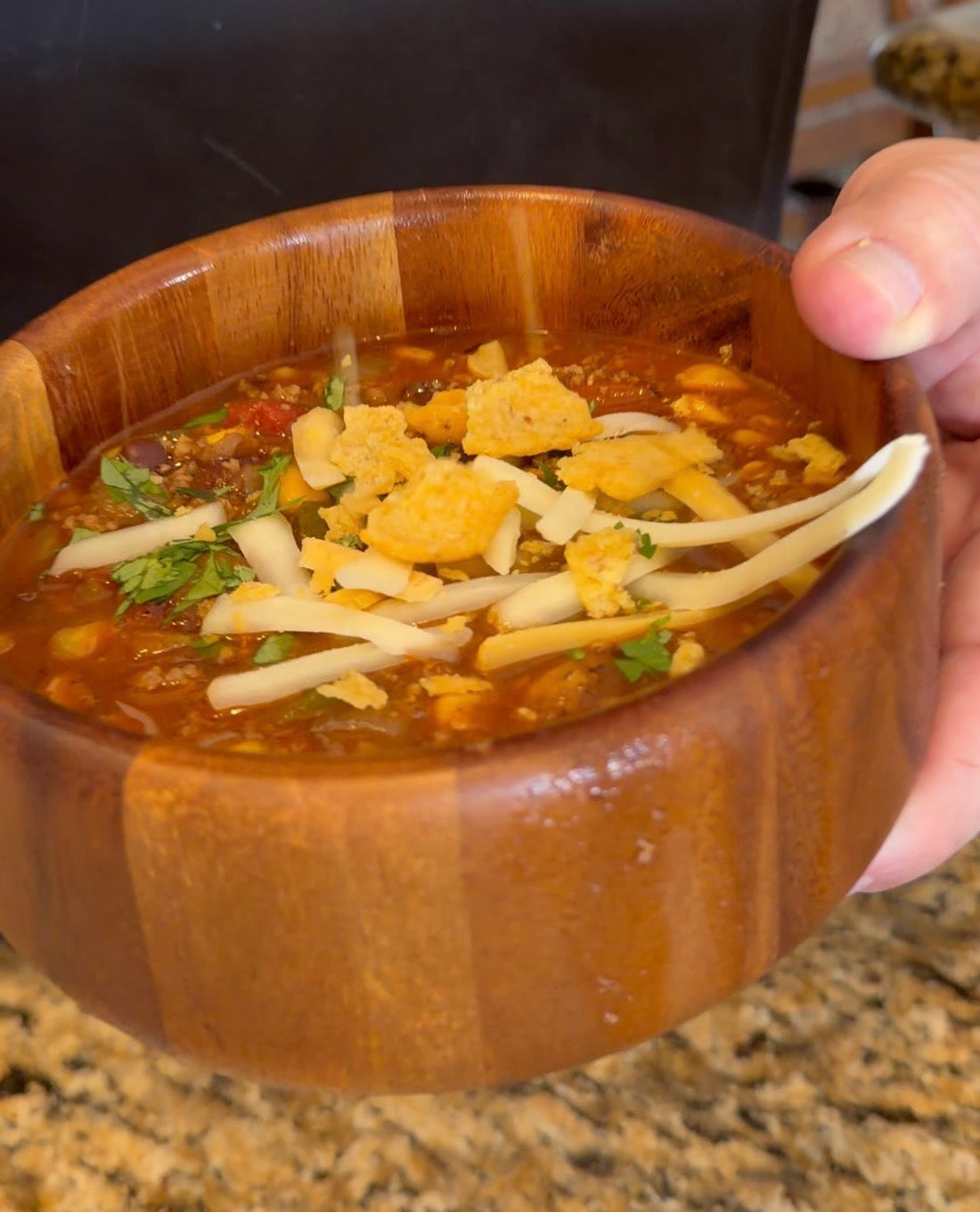 Ground beef taco soup with black beans and corn, topped with shredded cheese, cilantro and crushed corn chips in a wooden bowl.